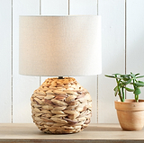 A woven natural fiber table lamp with a cylindrical drum shade and a round braided base, shown on a wooden surface next to a small potted succulent against a white paneled wall.