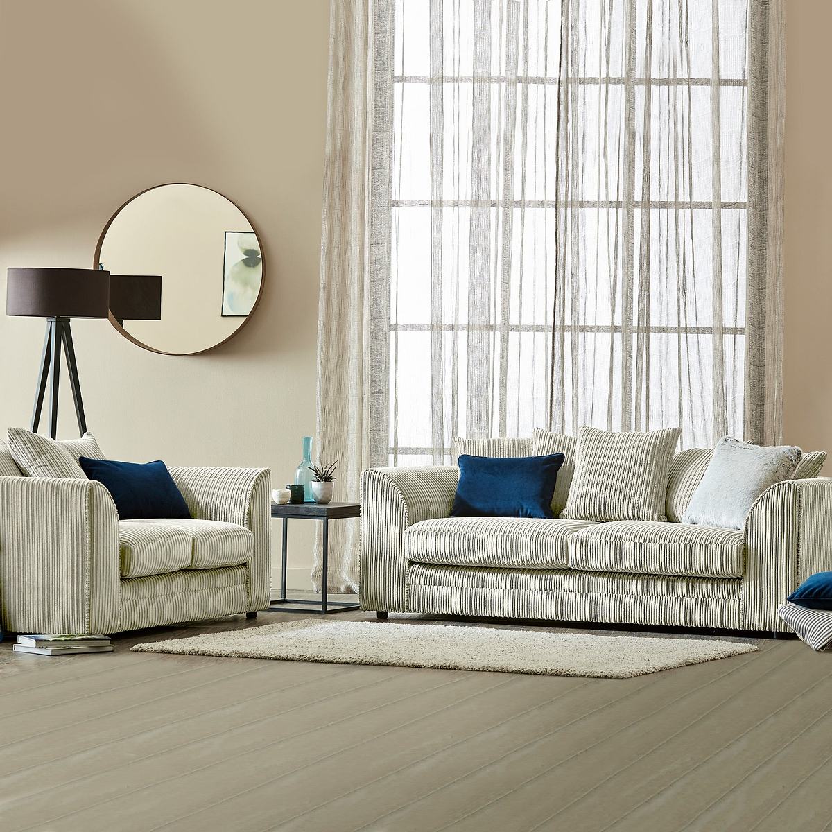 Striped sofa and armchair with blue and white cushions, a floor lamp, and round mirror, in a neutral-toned living room with sheer curtained window and wooden floor.
