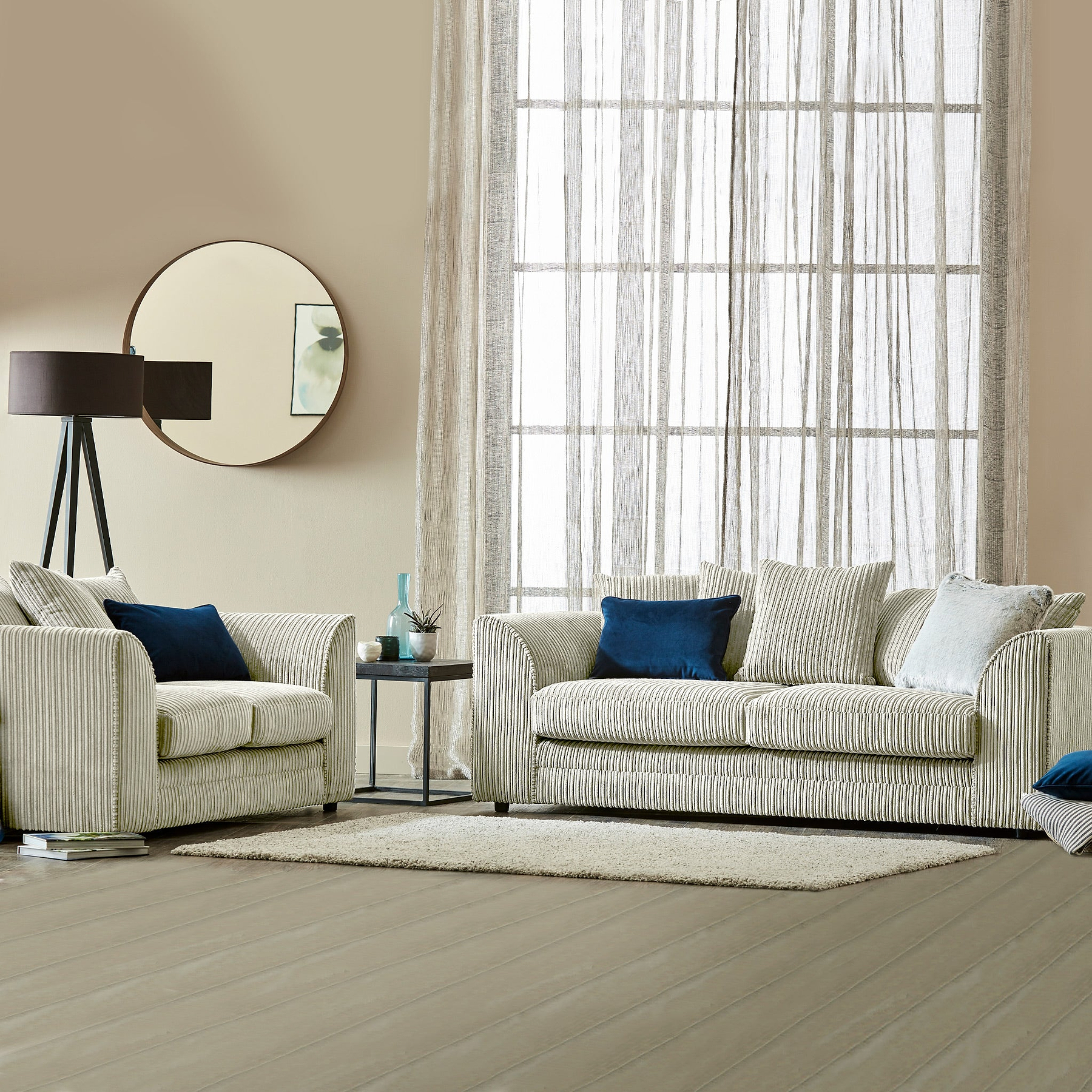 Striped sofa and armchair with blue and white cushions, a floor lamp, and round mirror, in a neutral-toned living room with sheer curtained window and wooden floor.