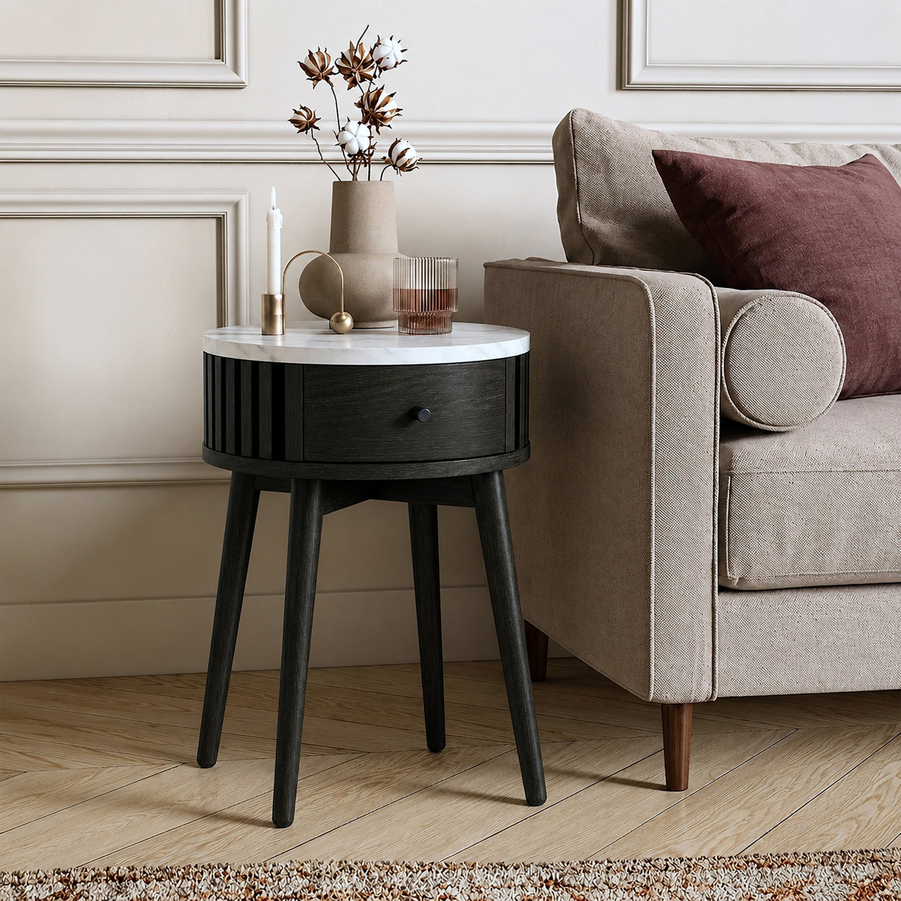 A modern Shorwell Black & Marble 1 Drawer Side Table with a white marble top and a dark charcoal black drawer and legs, styled next to a beige armchair in a living room setting.