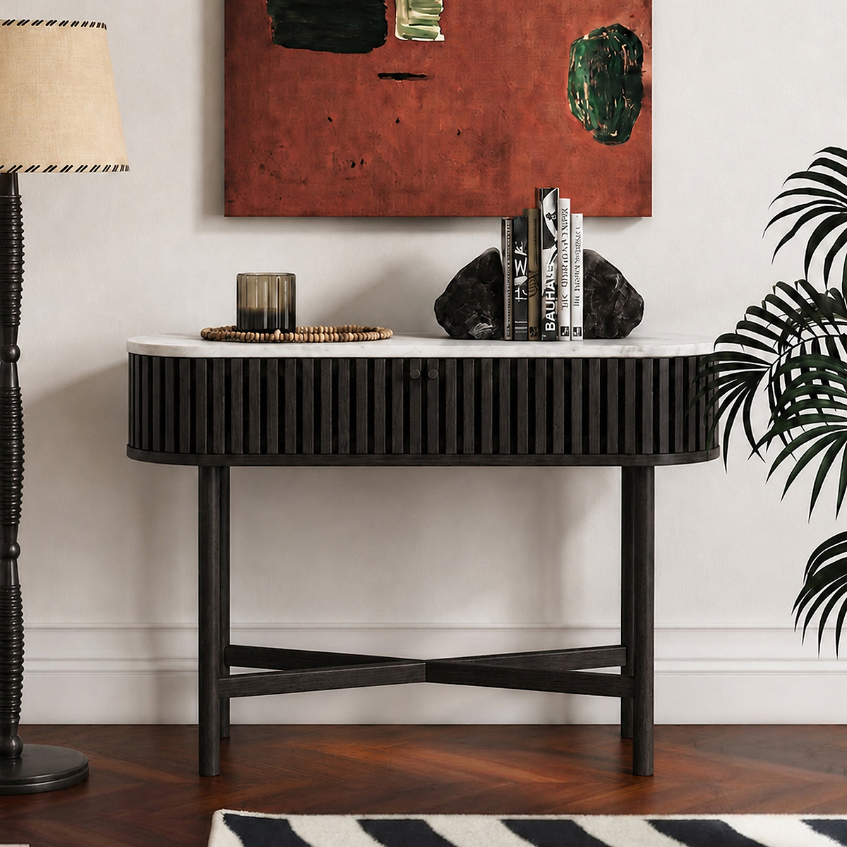 A modern Shorwell Black & Marble Oval Console Table  with a black slatted frame, white marble top, and black legs, styled in a living room setting with books, a candle, and decorative rocks.