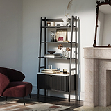 A Shorwell Black & Marble Bookcase with a slatted lower cabinet door, styled with books, vases, and decor in a modern living room next to a burgundy armchair and a stone fireplace.