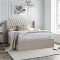 A natural upholstered bed with a tall, tufted headboard is set against a paneled wall on herringbone-patterned wooden flooring.
