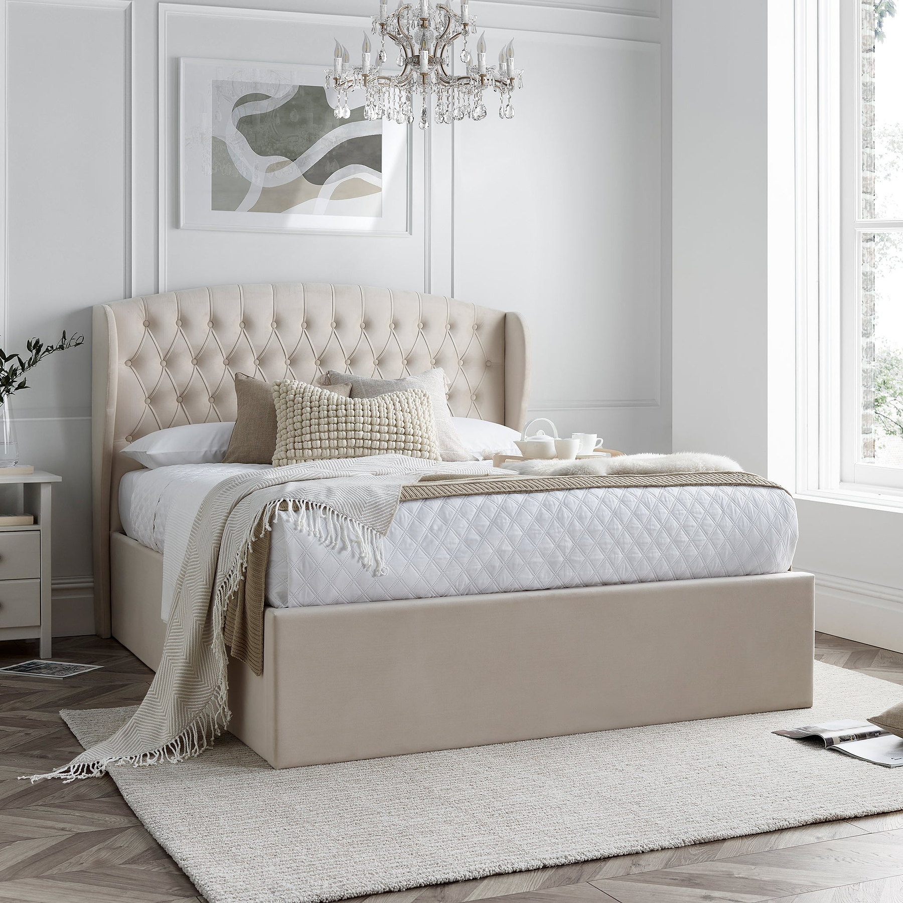 A natural upholstered bed with a tall, tufted headboard is set against a paneled wall on herringbone-patterned wooden flooring.
