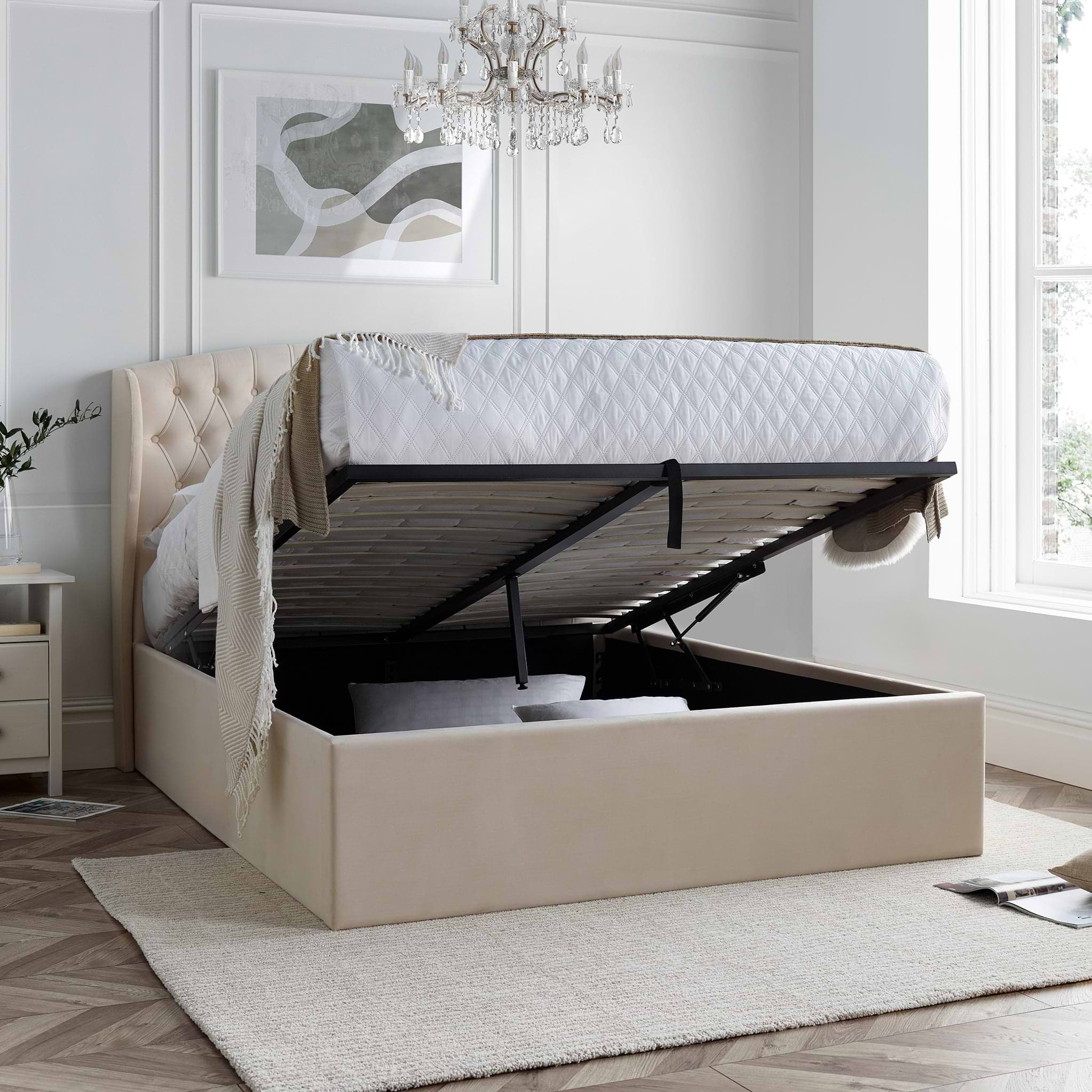 A natural upholstered ottoman bed with a lifted mattress reveals storage underneath, positioned against a paneled wall on a herringbone wood floor.