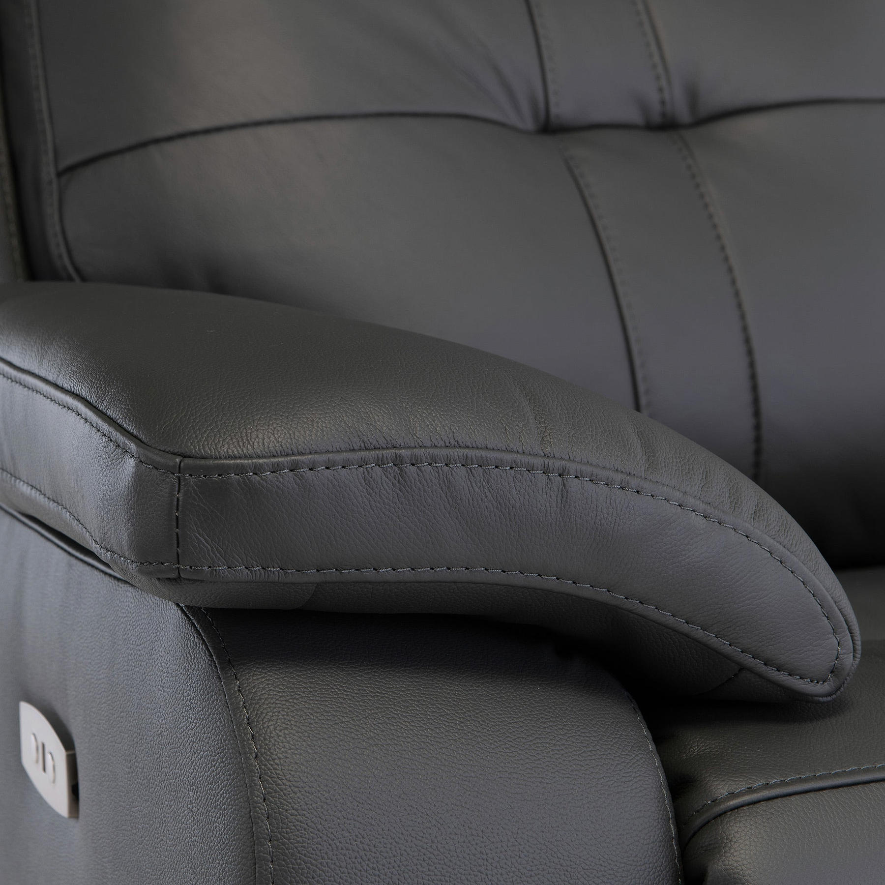 A black leather armchair with visible stitching, focused on the curved armrest, against a dark, indistinct background.