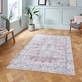 A patterned rug lies on a herringbone parquet floor in a room with paneled walls, a potted plant, bench, books, and a pastel-colored chair.