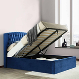A blue upholstered storage bed with the mattress lifted up, revealing an open storage space beneath, set against a paneled wall on a herringbone floor.
