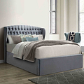 A gray upholstered bed with a tall, tufted headboard is set against a paneled wall on herringbone-patterned wooden flooring.