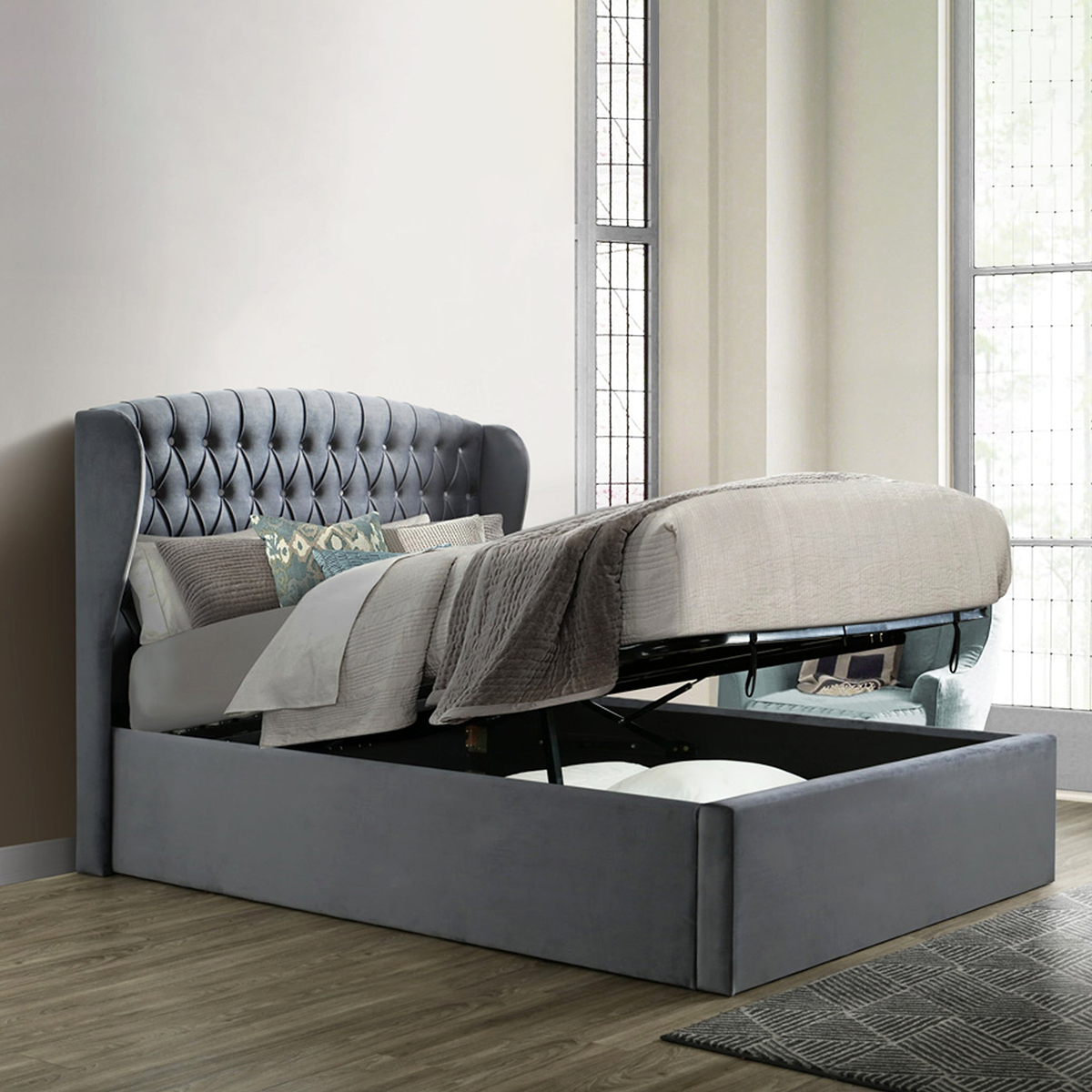 An open gray storage bed with a lifted mattress reveals a spacious compartment underneath, situated against a paneled wall on a herringbone-patterned wooden floor.