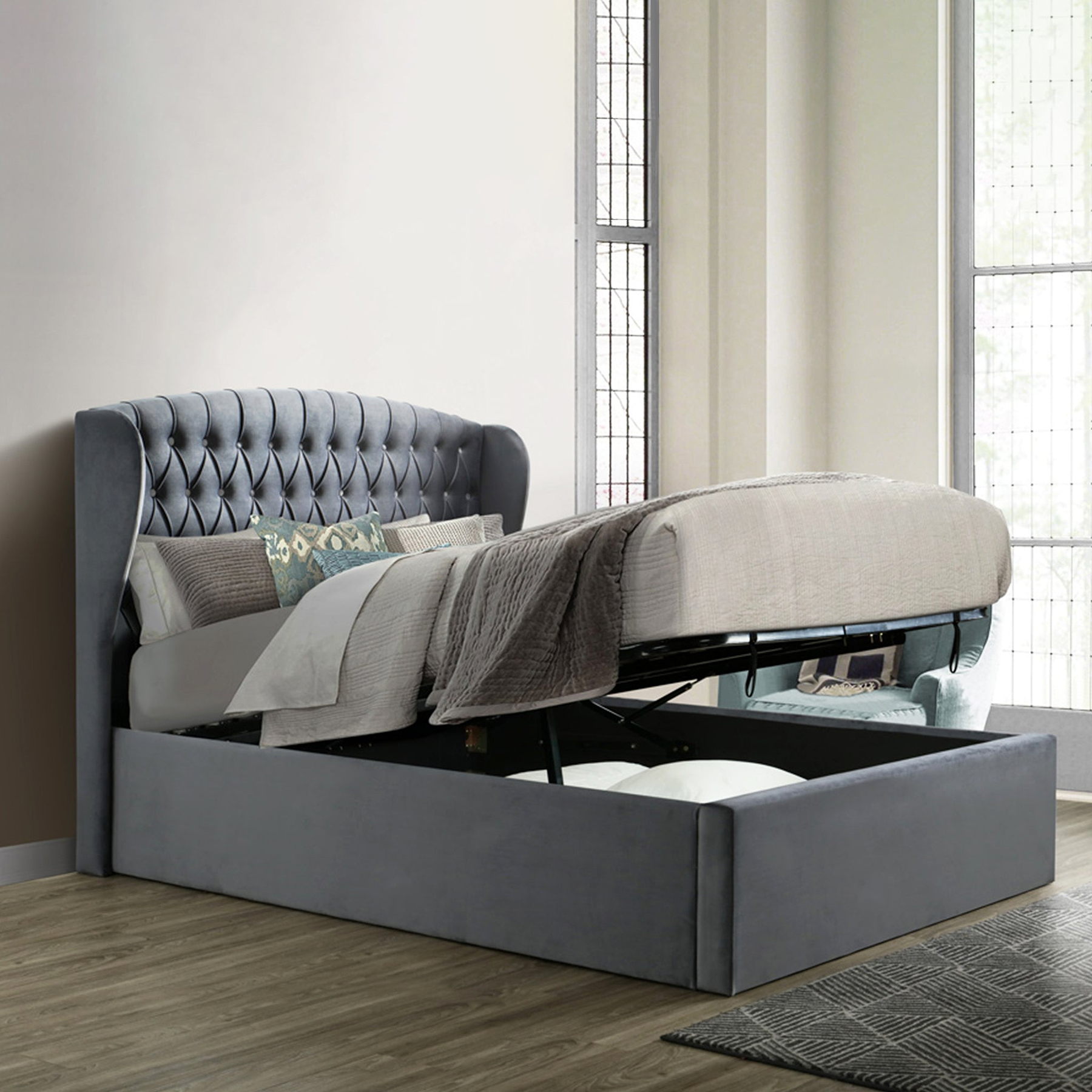 An open gray storage bed with a lifted mattress reveals a spacious compartment underneath, situated against a paneled wall on a herringbone-patterned wooden floor.