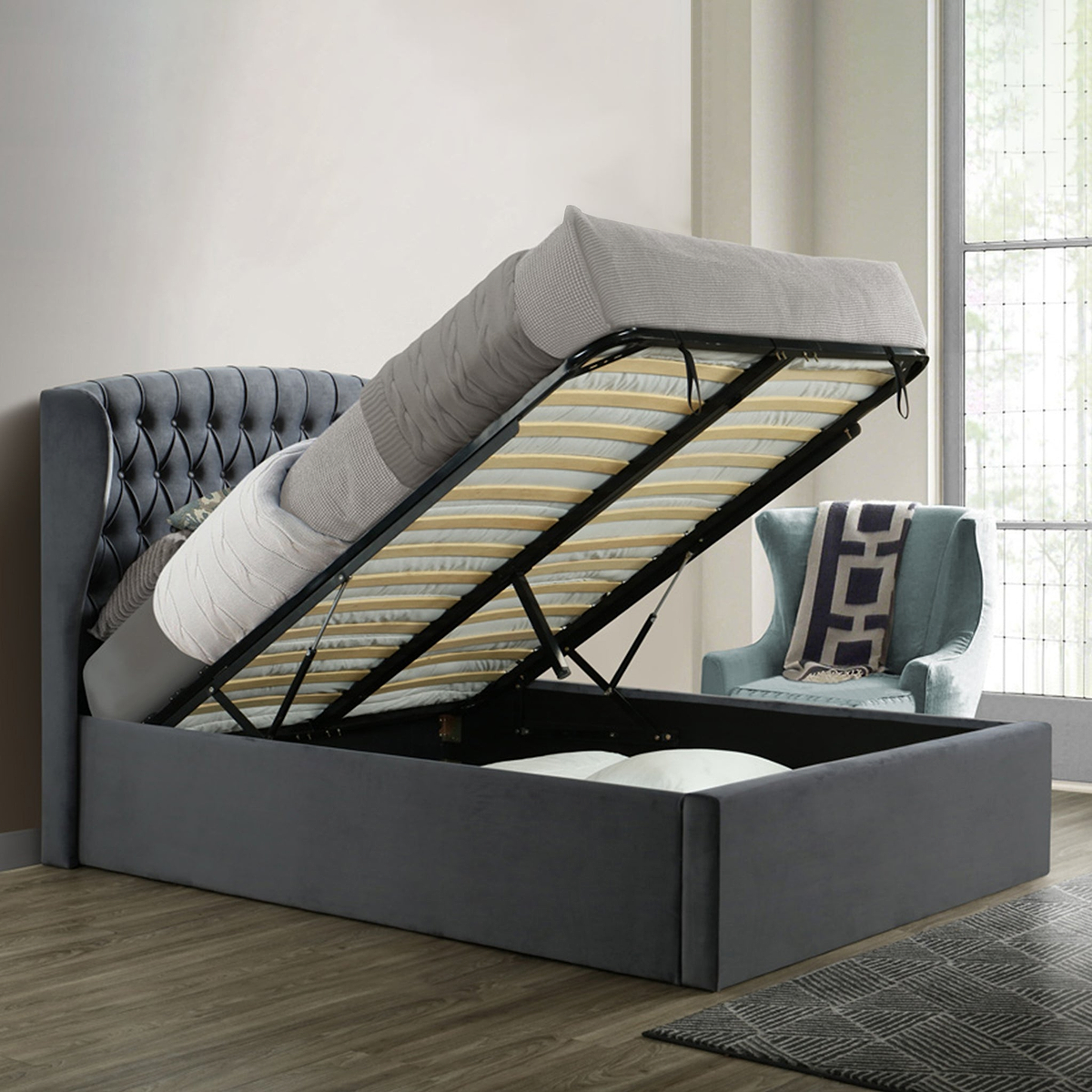 A gray upholstered bed with a lifted mattress reveals storage underneath, positioned against a paneled wall on a herringbone wood floor.