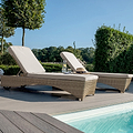 Two sun loungers by a pool with cushions, a hat, and a glass of water, set against a backdrop of trees and shrubs in a garden during daytime.