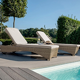 Two sun loungers by a pool with cushions, a hat, and a glass of water, set against a backdrop of trees and shrubs in a garden during daytime.