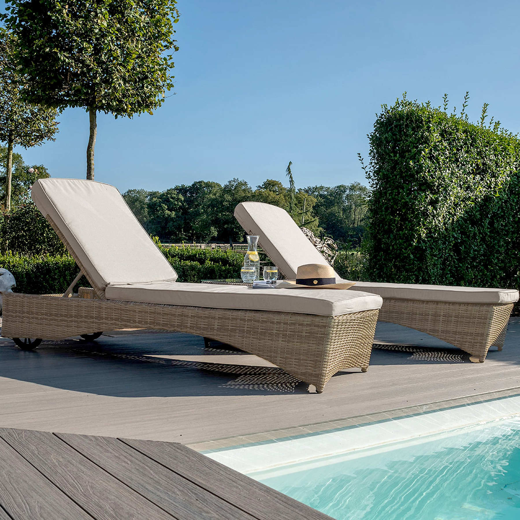 Two sun loungers by a pool with cushions, a hat, and a glass of water, set against a backdrop of trees and shrubs in a garden during daytime.