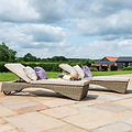 Two wicker sun loungers with beige cushions and pillows are set on a paved patio, under a clear blue sky with puffy clouds, surrounded by greenery.