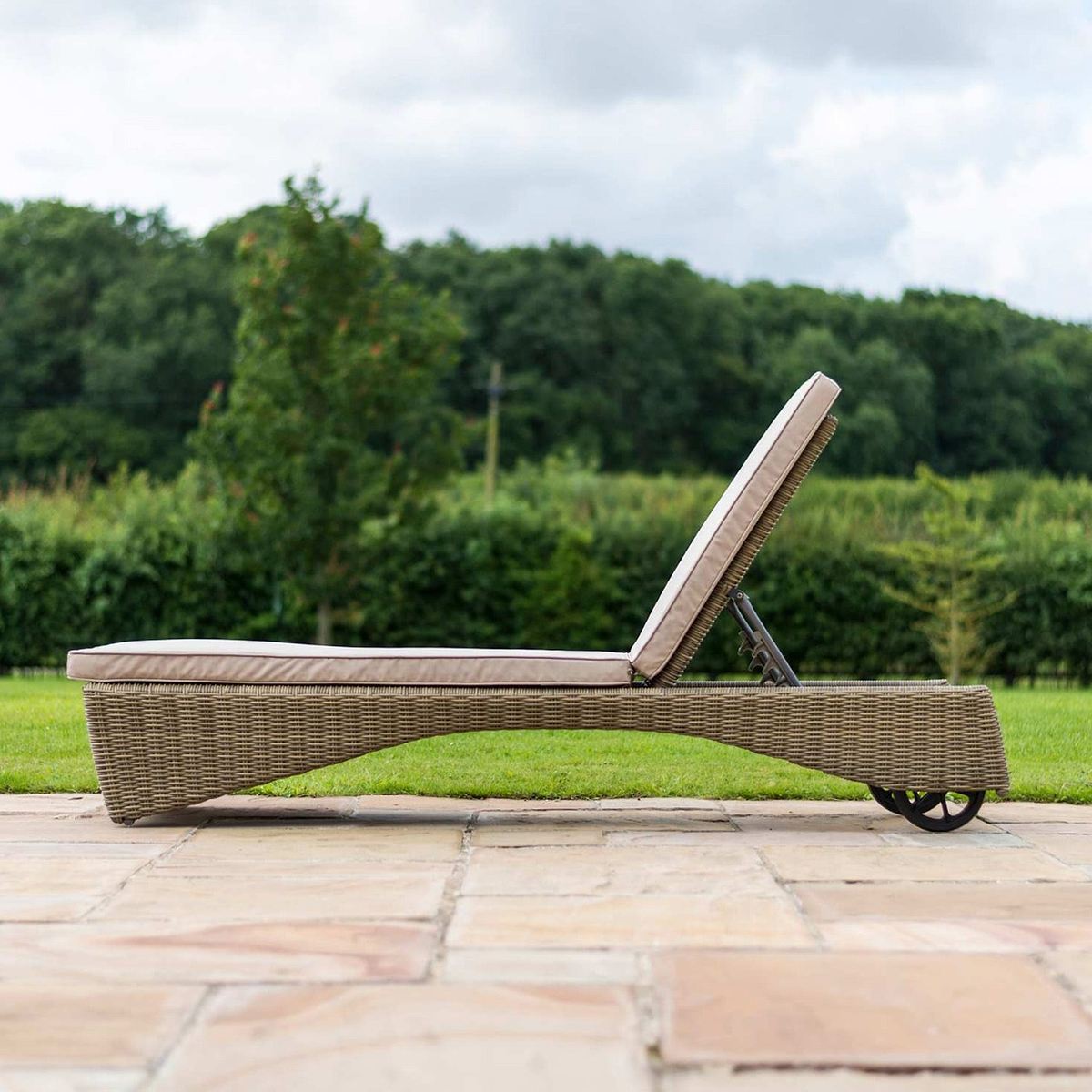 A wicker sun lounger with a beige cushion positioned on a paved patio, backed by a lush green lawn and trees.