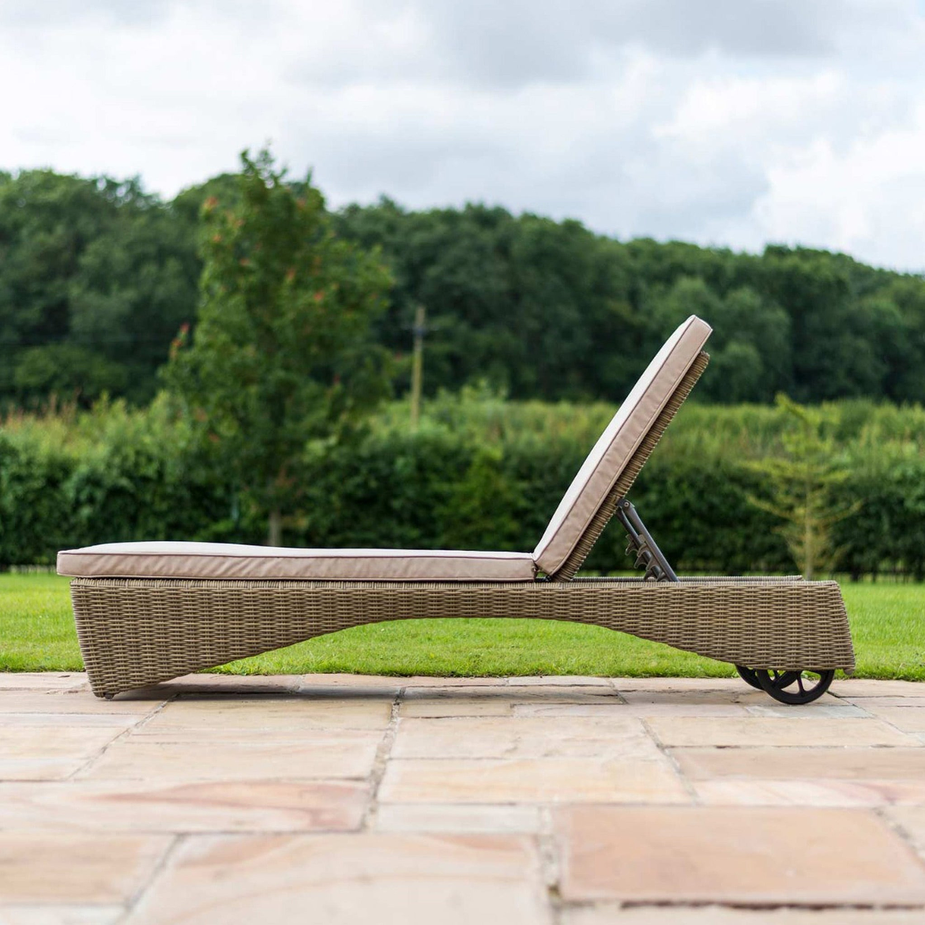 A wicker sun lounger with a beige cushion positioned on a paved patio, backed by a lush green lawn and trees.