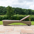 A wicker sun lounger with a cushion rests on a stone patio, amidst a lush green lawn and trees in the background.