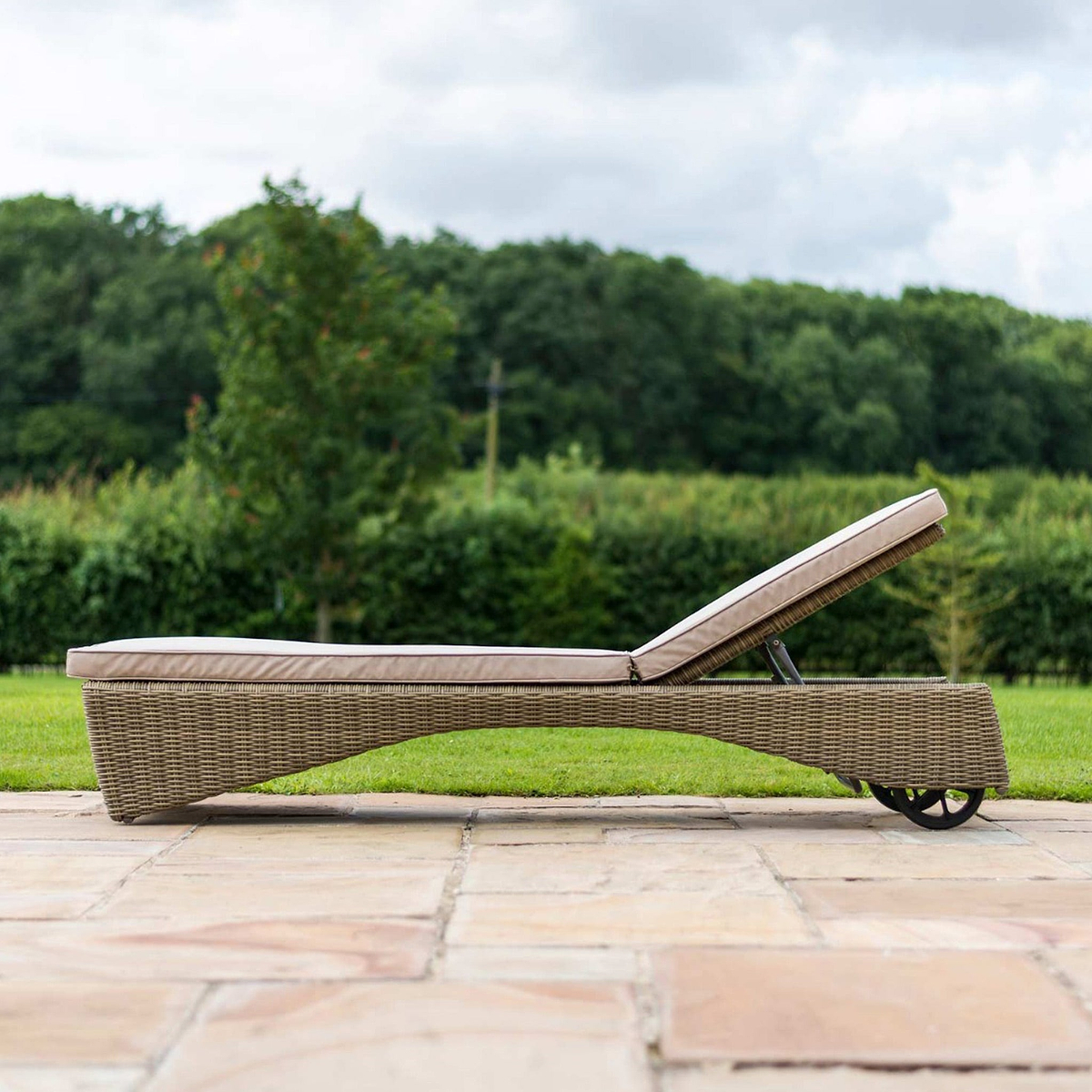 A wicker sun lounger with a cushion rests on a stone patio, amidst a lush green lawn and trees in the background.