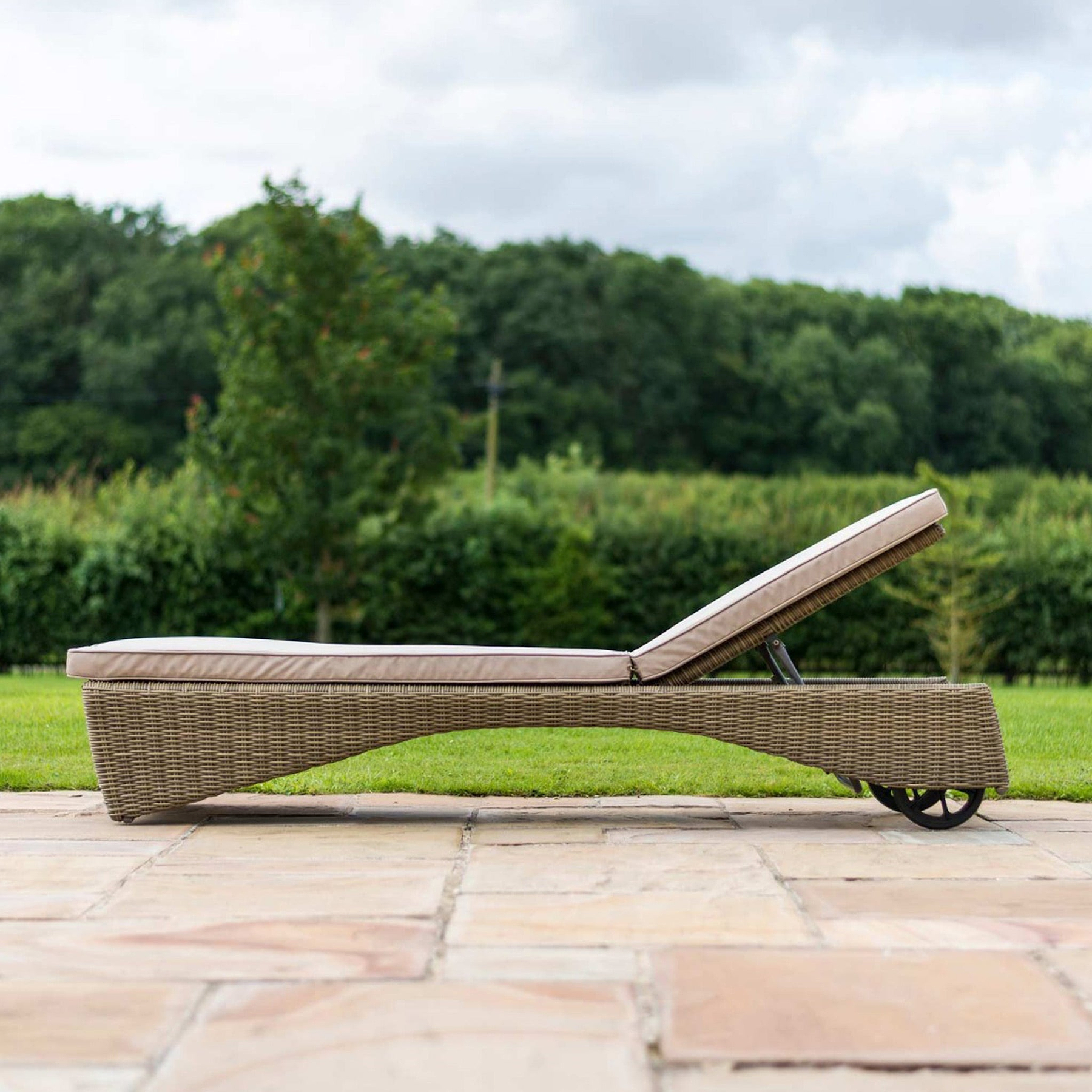 A wicker sun lounger with a cushion rests on a stone patio, amidst a lush green lawn and trees in the background.