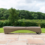 A curved, wicker sun lounger with a cushion rests on a paved patio, with lush greenery in the background.