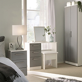 A serene bedroom space with a dressing table and mirror, flanked by a wardrobe and a bedside drawer with a lamp. A window with sheer curtains brightens the room.