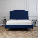 A blue upholstered bed with a white mattress stands stationary against a white paneled wall, with a black side table nearby, set on a herringbone wood floor.