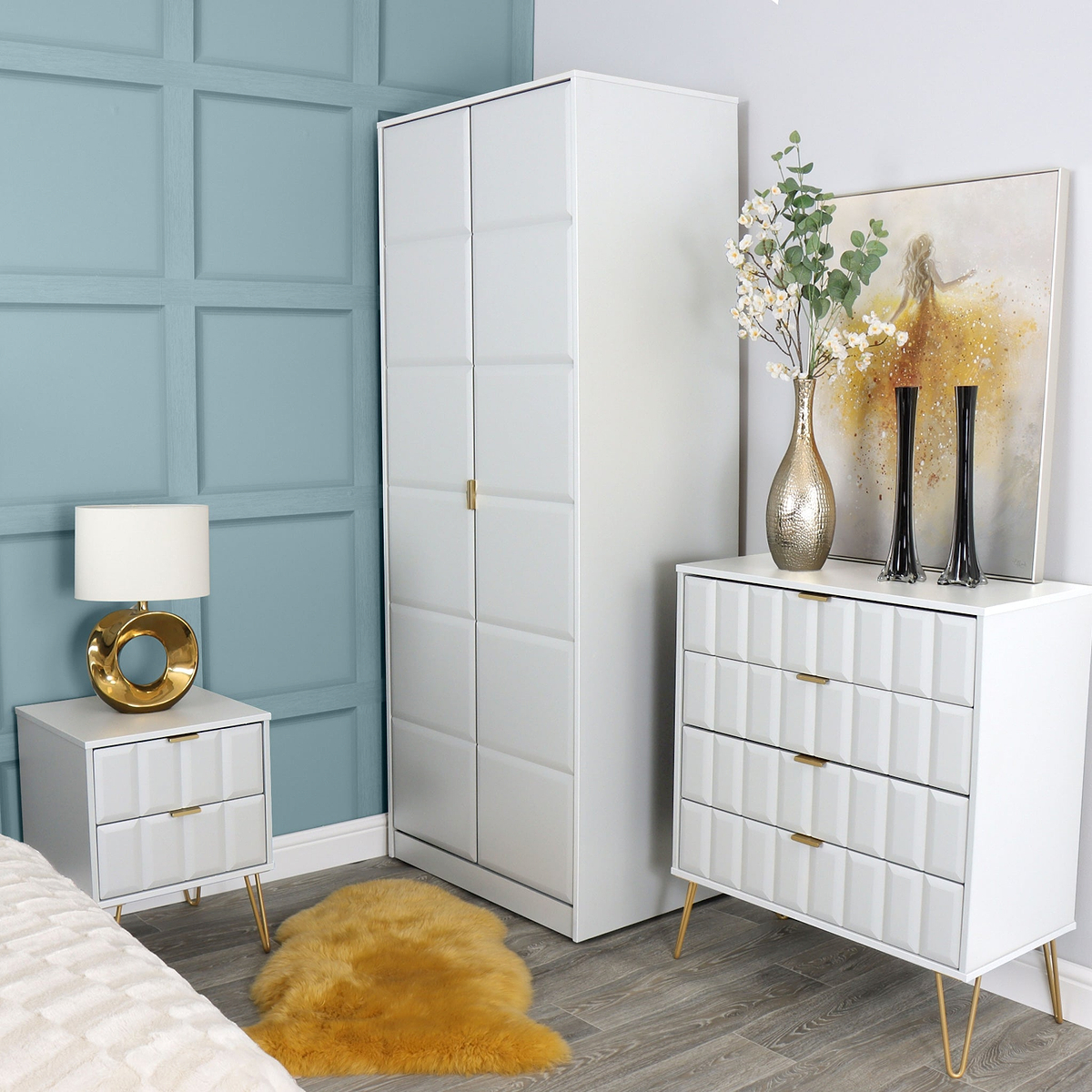A modern bedroom corner with a white wardrobe and dresser against a blue paneled wall, accessorized with a gold and white lamp, a vase with branches, and dark candlesticks. A yellow rug is on the floor.