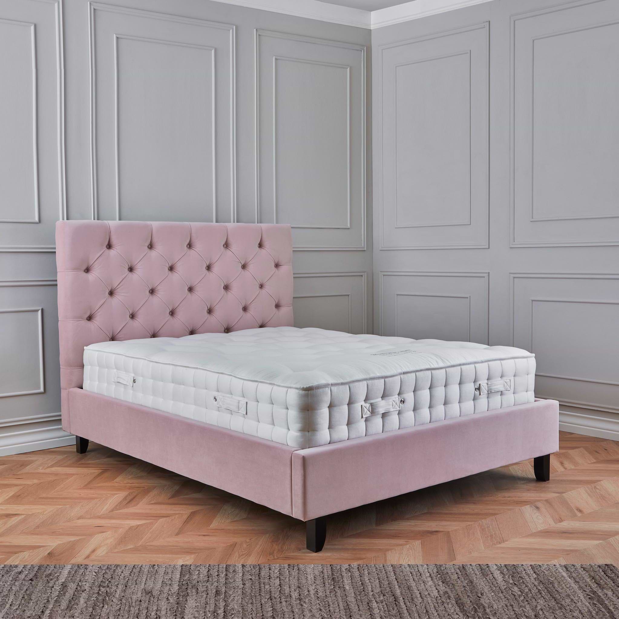 A pink, tufted headboard rests behind a plush white mattress on a bed frame, positioned in a room with elegant grey paneled walls and wooden flooring.