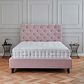 A plush pink bed with a tufted headboard sits in a room with classic paneled walls and a herringbone parquet floor. A small side table and decorative items accompany it.