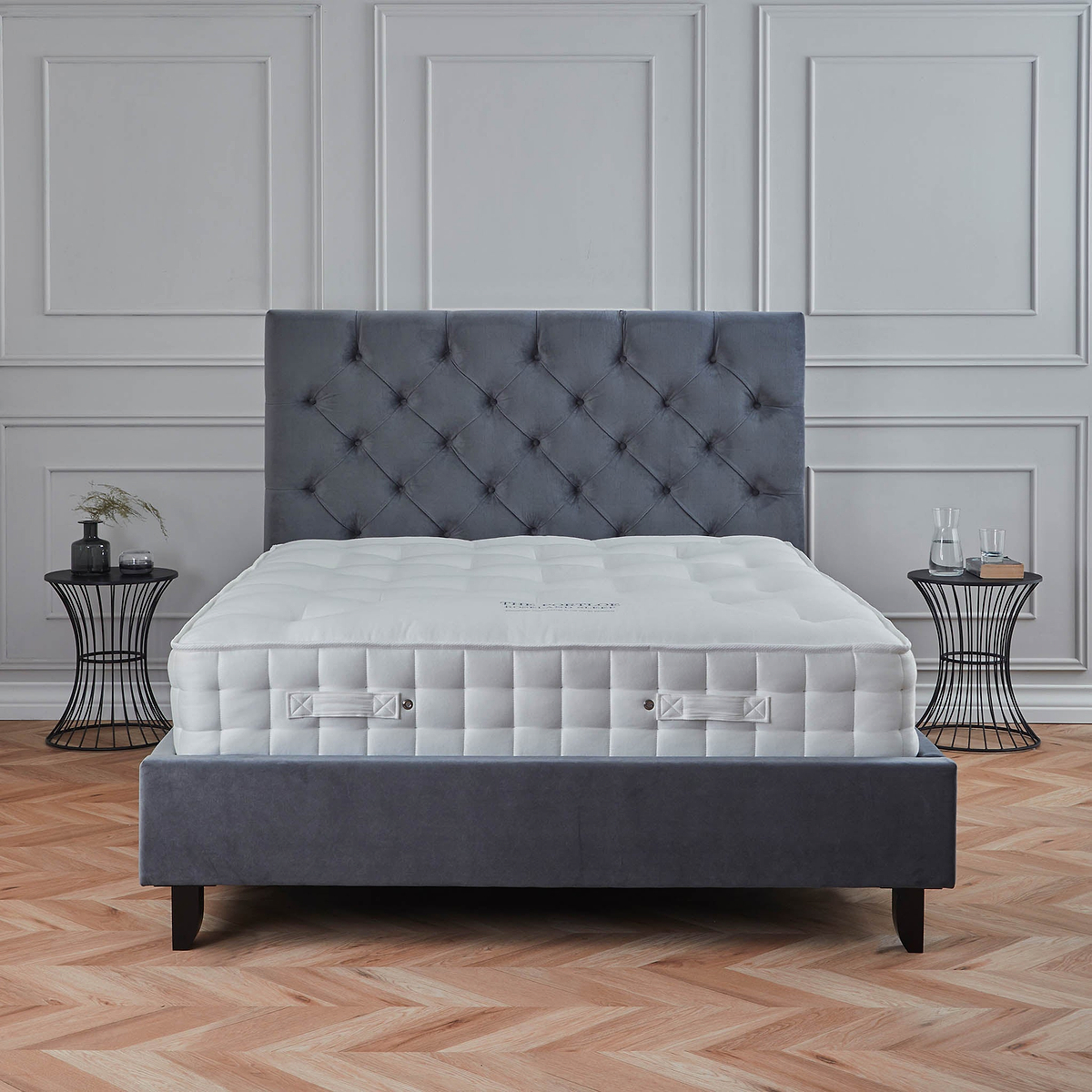 A gray upholstered bed with a quilted headboard and mattress set in a room with paneled walls and herringbone wood flooring, accompanied by a wireframe side table.