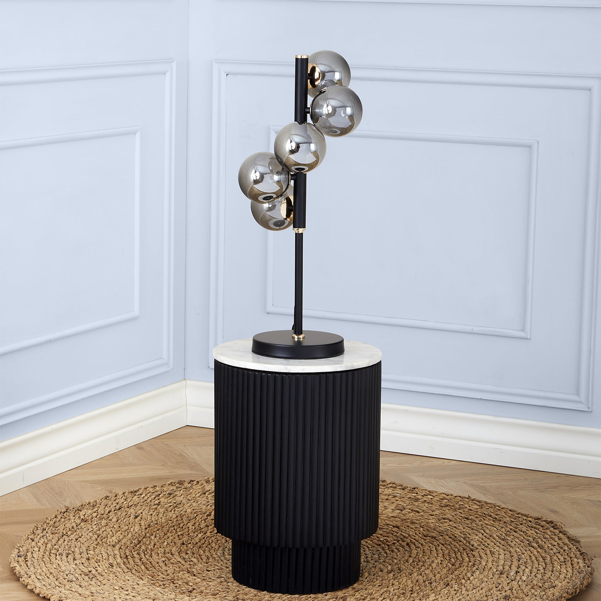 A sleek, modern floor lamp with five spherical, smoky glass shades mounted at varying heights on a vertical black pole, set against a pale blue paneled wall and standing on a ribbed cylindrical base atop a round jute rug.