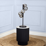 A sleek, modern floor lamp with five spherical, smoky glass shades mounted at varying heights on a vertical black pole, set against a pale blue paneled wall and standing on a ribbed cylindrical base atop a round jute rug.