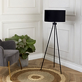 A tall, black tripod floor lamp stands illuminated in a classy room, next to a grey armchair and a potted plant, over a circular woven rug on a wooden floor.