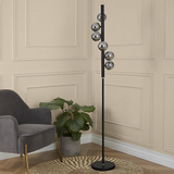 A black floor lamp with multiple spherical, silver shades stands in a room, next to a grey armchair and a potted plant on a woven rug, against a paneled wall.