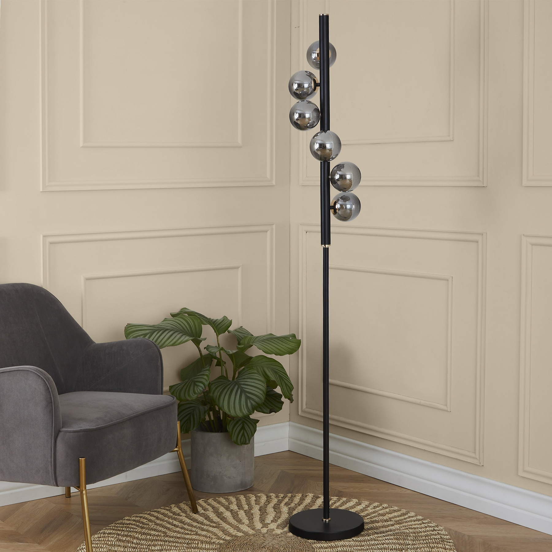 A black floor lamp with multiple spherical, silver shades stands in a room, next to a grey armchair and a potted plant on a woven rug, against a paneled wall.