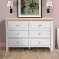 A white six-drawer wooden dresser sits against a pink wall, flanked by black wall lamps, under a framed painting of a Venetian canal scene.