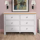 A white six-drawer wooden dresser sits against a pink wall, flanked by black wall lamps, under a framed painting of a Venetian canal scene.