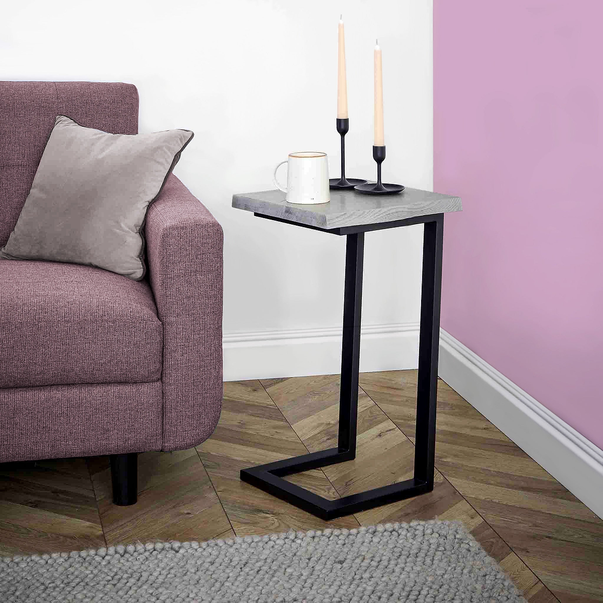 A side table with a mug and candles resting on it, situated next to a pink armchair, against a white and purple wall, above a grey carpet.