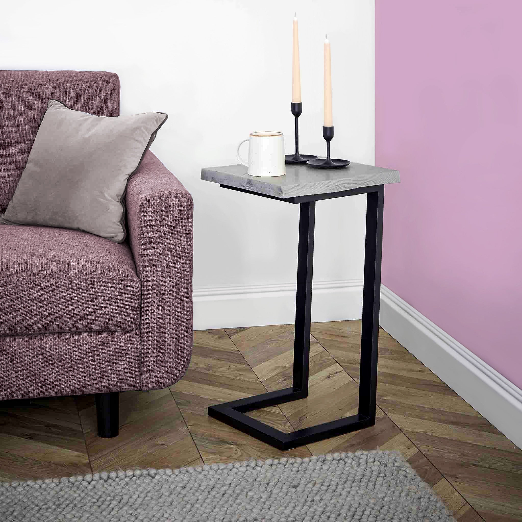 A side table with a mug and candles resting on it, situated next to a pink armchair, against a white and purple wall, above a grey carpet.