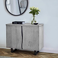 A wooden cabinet stands against a wall, atop which rests a book labeled "CHANEL," a vase with white flowers, and a small bowl, near a round mirror.