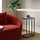 A red velvet sofa and a slender, black metal side table holding a book and black cup in a room with wood flooring and a potted plant.