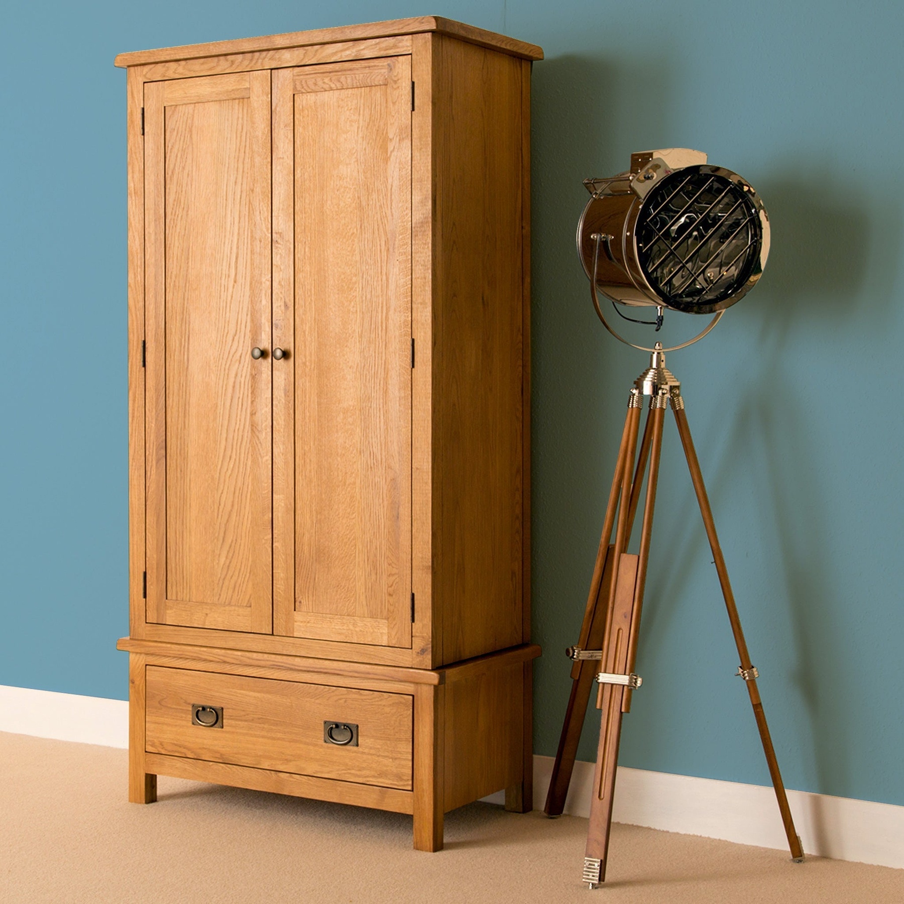 A wooden armoire stands next to a tripod-based spotlight lamp against a teal wall on a beige carpet.
