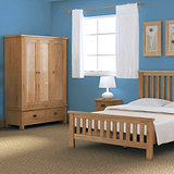 A wooden bed and wardrobe furnish a bedroom with blue walls, white curtains on a window, and a lamp on a bedside table.
