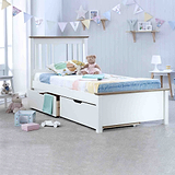A white single bed with colorful bedding sits in a bright child's bedroom. Plush toys are on and around the bed, with a bookshelf and decorations nearby.