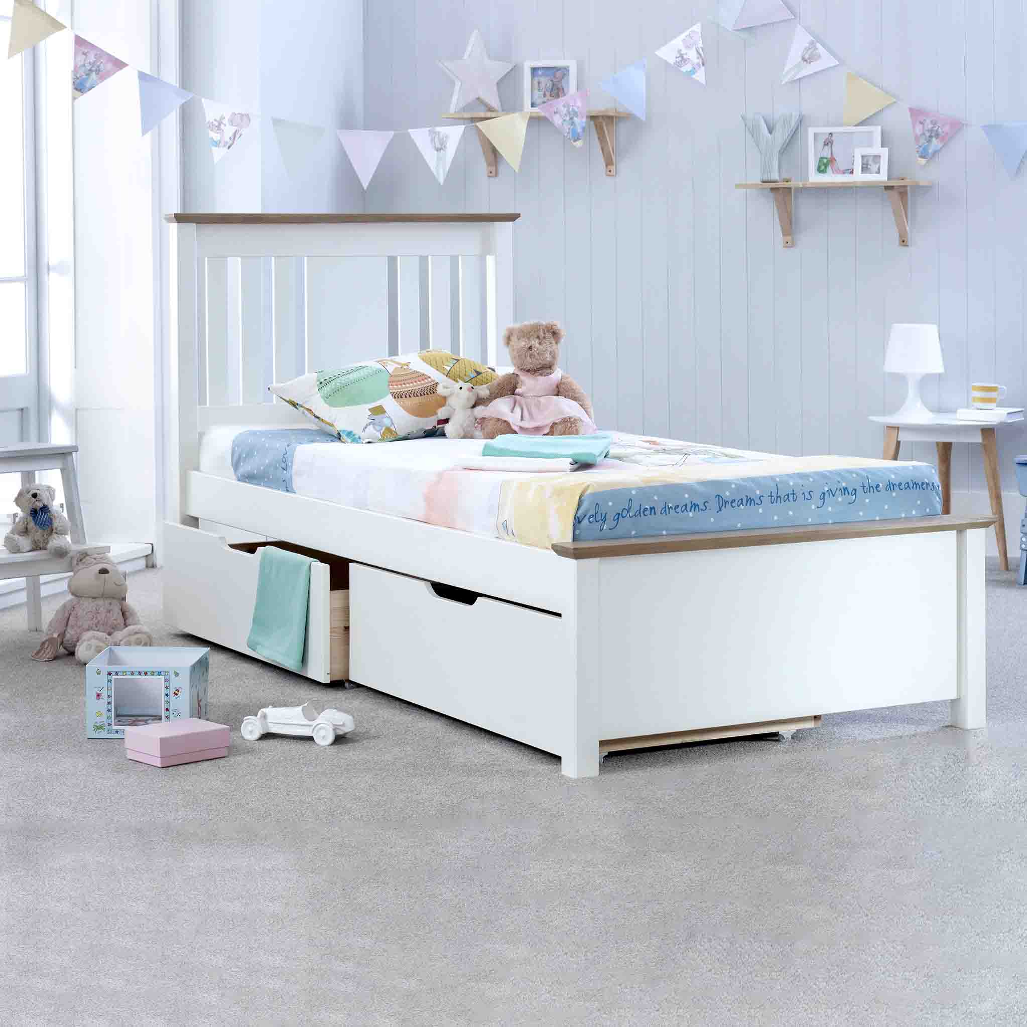 A white single bed with colorful bedding sits in a bright child's bedroom. Plush toys are on and around the bed, with a bookshelf and decorations nearby.