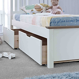 A white children's bed with storage drawers underneath sits adorned with pillows and stuffed toys, in a tidy, bright room with a toy car on the floor.