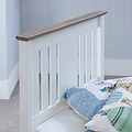 A white wooden bed frame with vertical slats stands against a grey wall, partially covered by a quilt with pastel patterns.