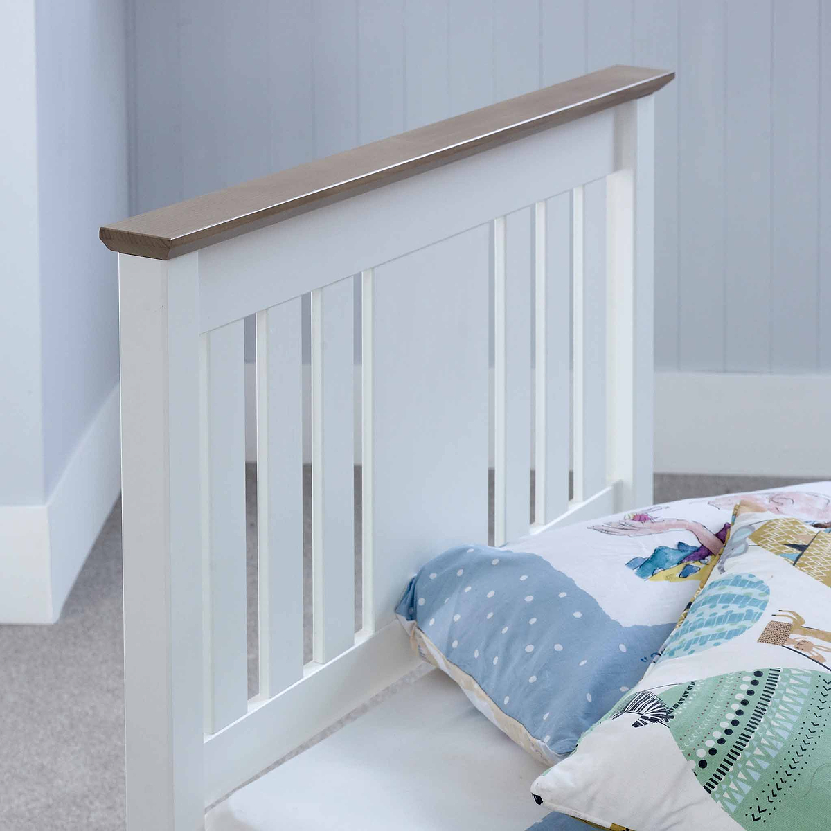 A white wooden bed frame with vertical slats stands against a grey wall, partially covered by a quilt with pastel patterns.